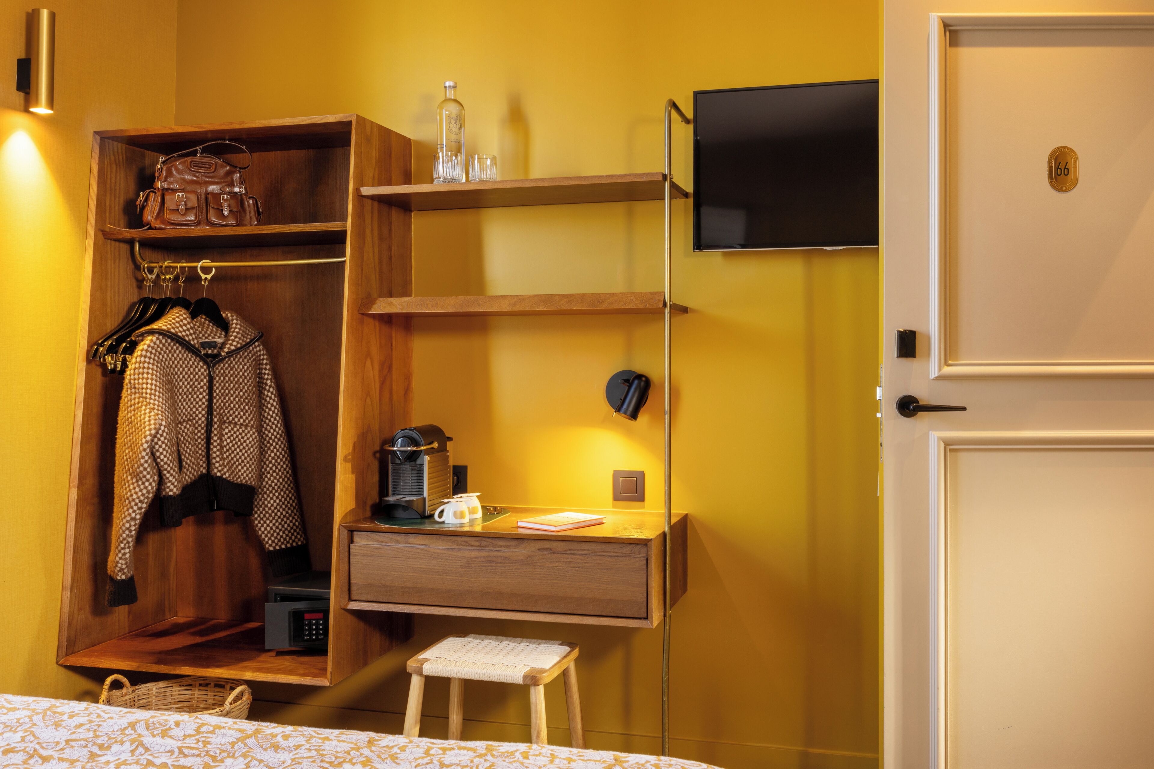 A hotel room with a wooden wardrobe, a desk with a coffee maker, a TV mounted on the wall, and a door with a number plate.