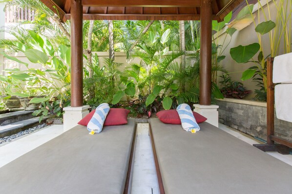 A covered outdoor seating area with two lounge chairs, red and striped cushions, and a variety of tropical plants.