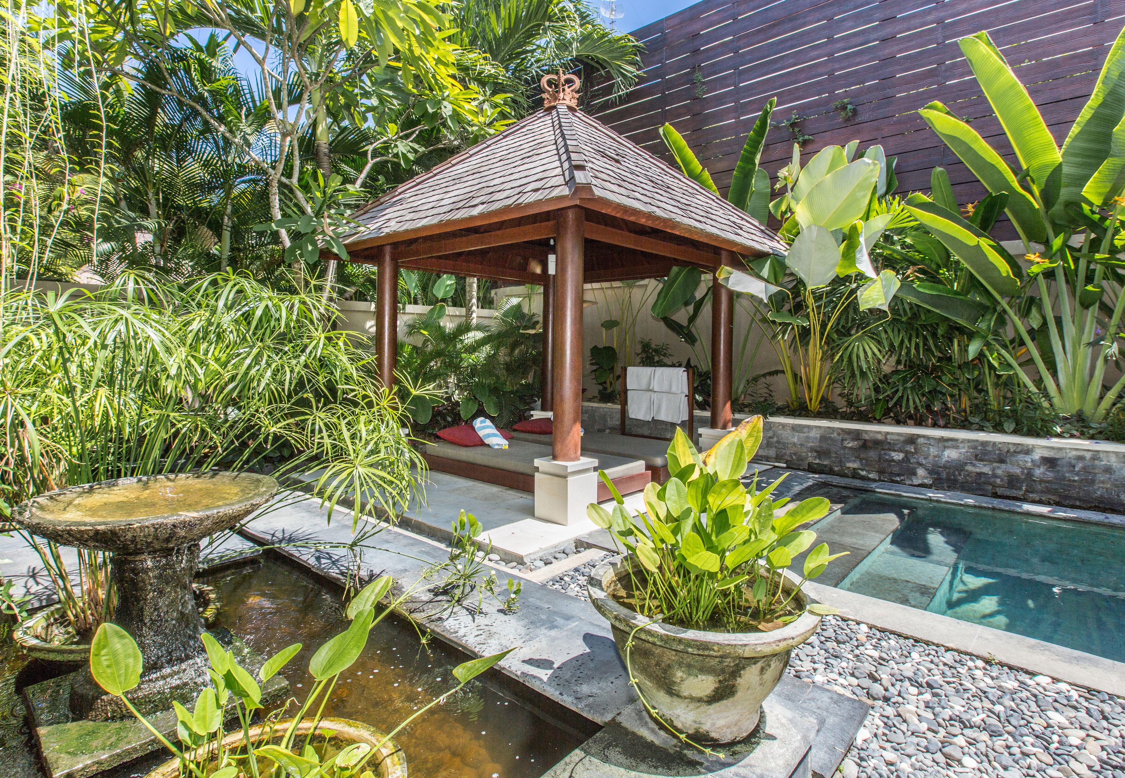 Ein Gartenlaube in einem Garten mit Pool, umgeben von verschiedenen Pflanzen und einem Holzzaun.