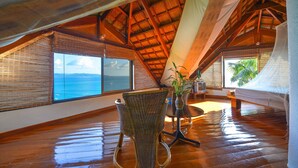 Une pièce avec un plafond en pente, un plancher en bois, une chaise en osier, une petite table avec un vase et un lit.