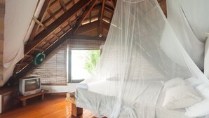 Une chambre avec un plafond en bois, un lit doté d’une moustiquaire, un téléviseur posé sur un meuble en bois et une fenêtre avec des stores.