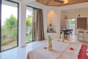 Habitación de hotel con un gran ventanal corredero a un jardín, una cama con cubrecama blanco y una zona de comedor con mesa y sillas.