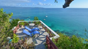 Une terrasse dotée de chaises longues bleues et d’une table, d’où l’on peut admirer une mer d’un bleu limpide et des bateaux.