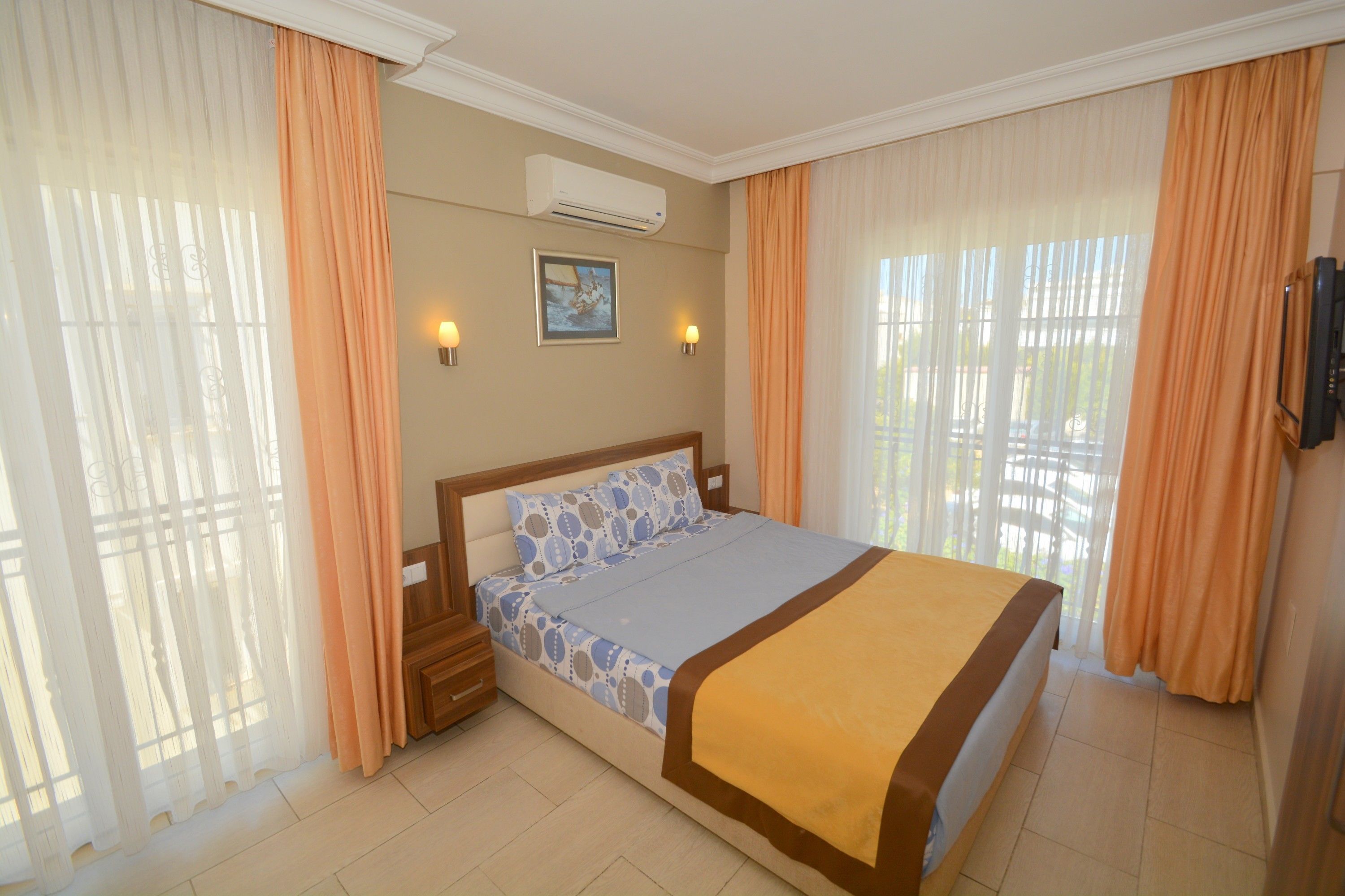 A hotel room with a bed, curtains, a wall-mounted air conditioner, a framed picture, and a television.