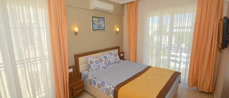 A hotel room with a bed, curtains, a wall-mounted air conditioner, a framed picture, and a television.