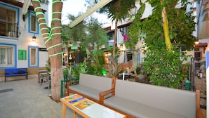 Un patio interior con zonas de estar, incluyendo un banco y una mesa, rodeado de diversas plantas y árboles.