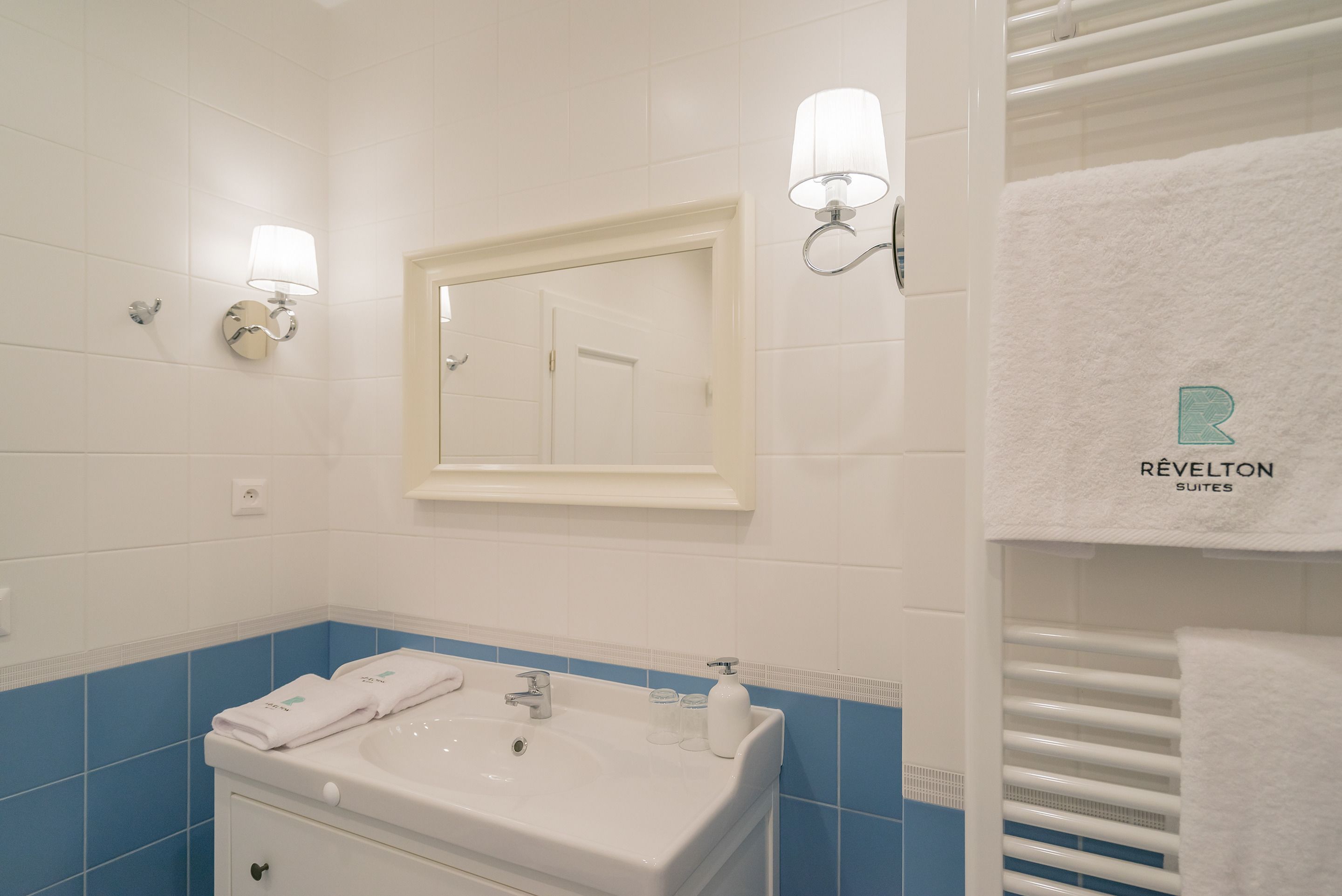 Een badkamer met een wit wastafelmeubel, blauwe tegels, een spiegel, twee wandlampen en een handdoekrek met een witte handdoek.
