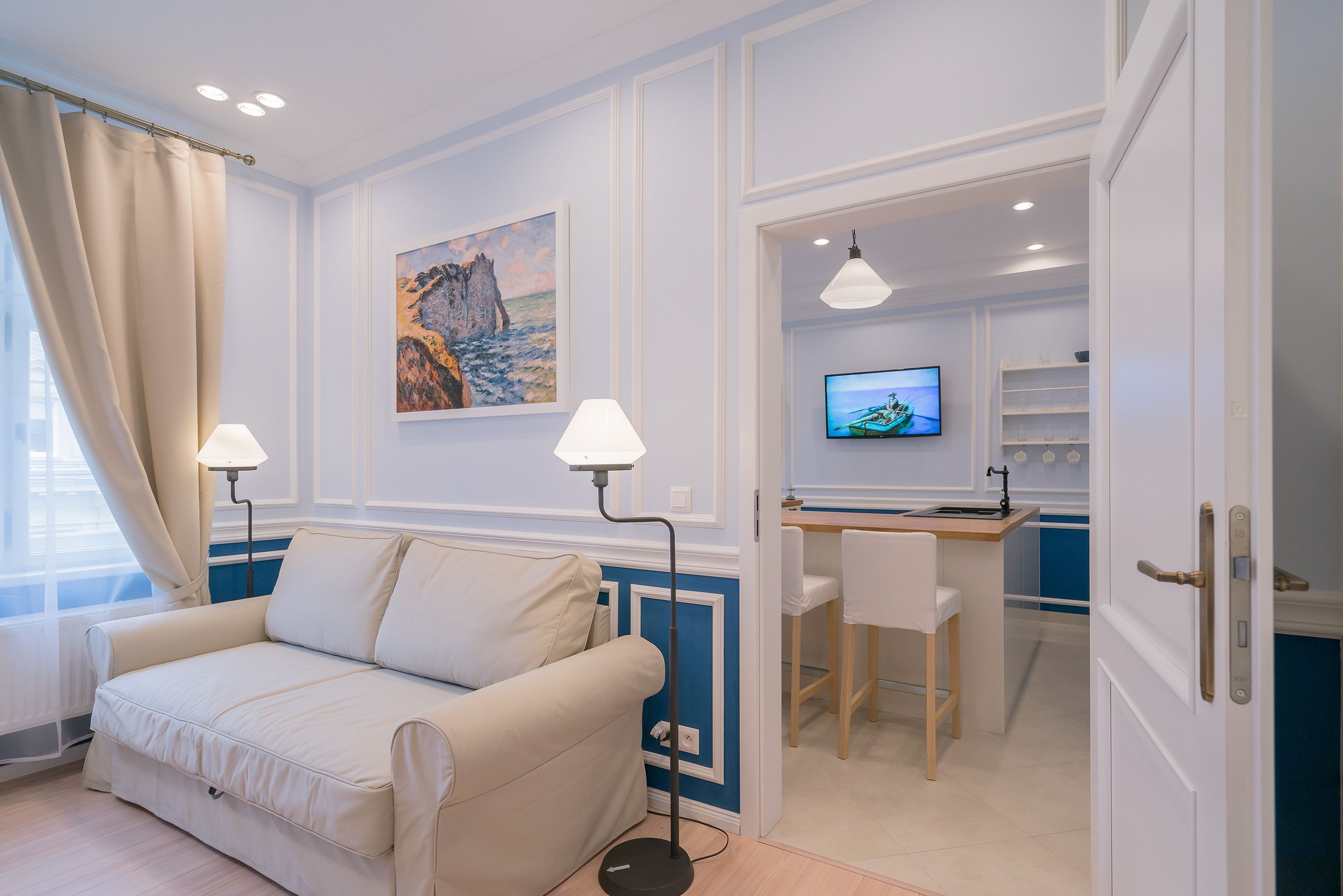 Een kamer met een beige bank, een bureau met stoel, een televisie en een schilderij aan de muur.