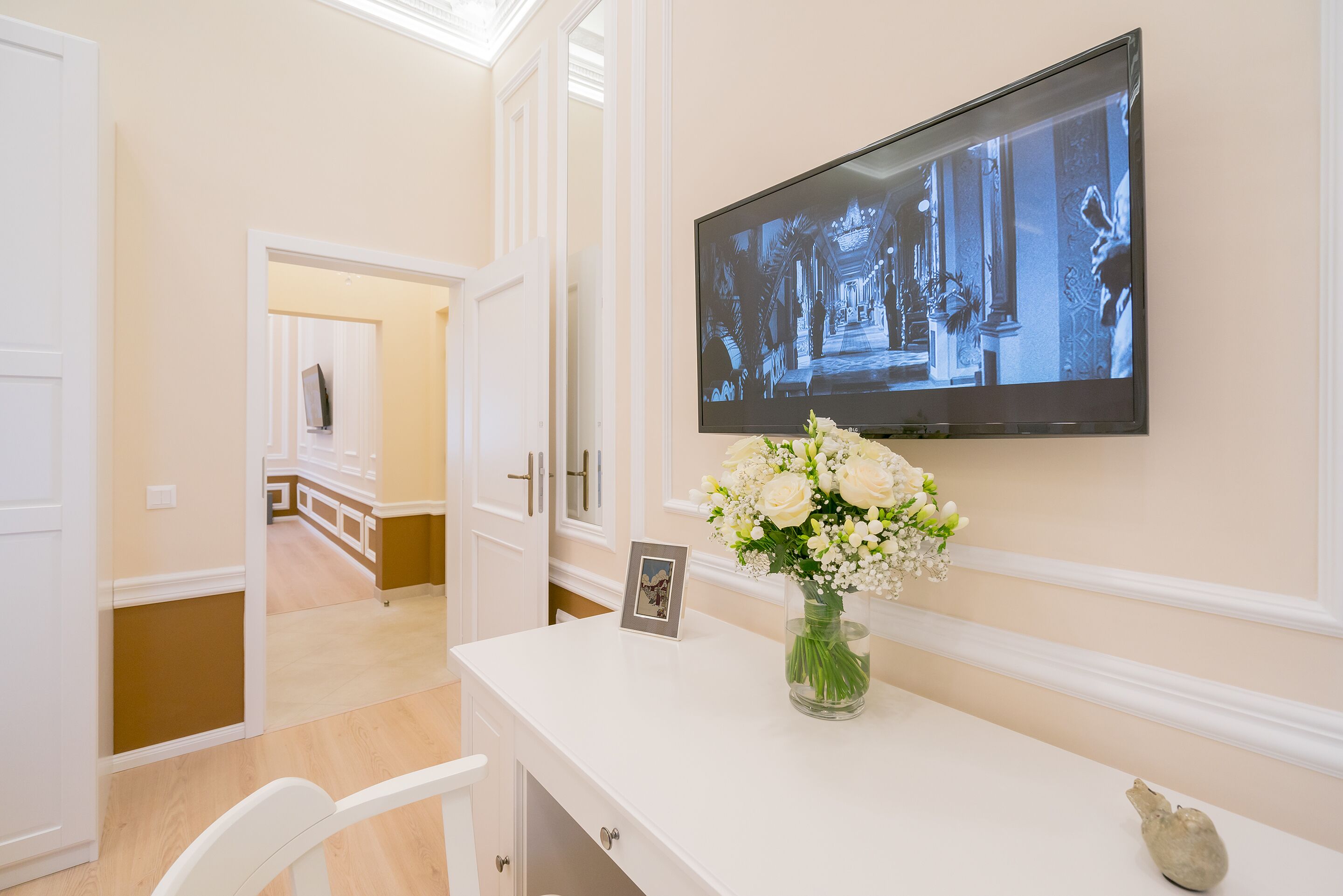 A hotel room with a white console table, a flat-screen TV mounted on the wall, a vase with flowers, and a framed photo.