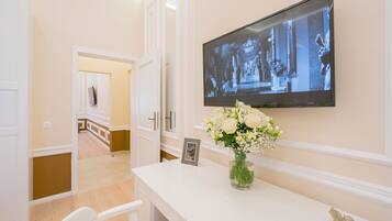 A hotel room with a white console table, a flat-screen TV mounted on the wall, a vase with flowers, and a framed photo.