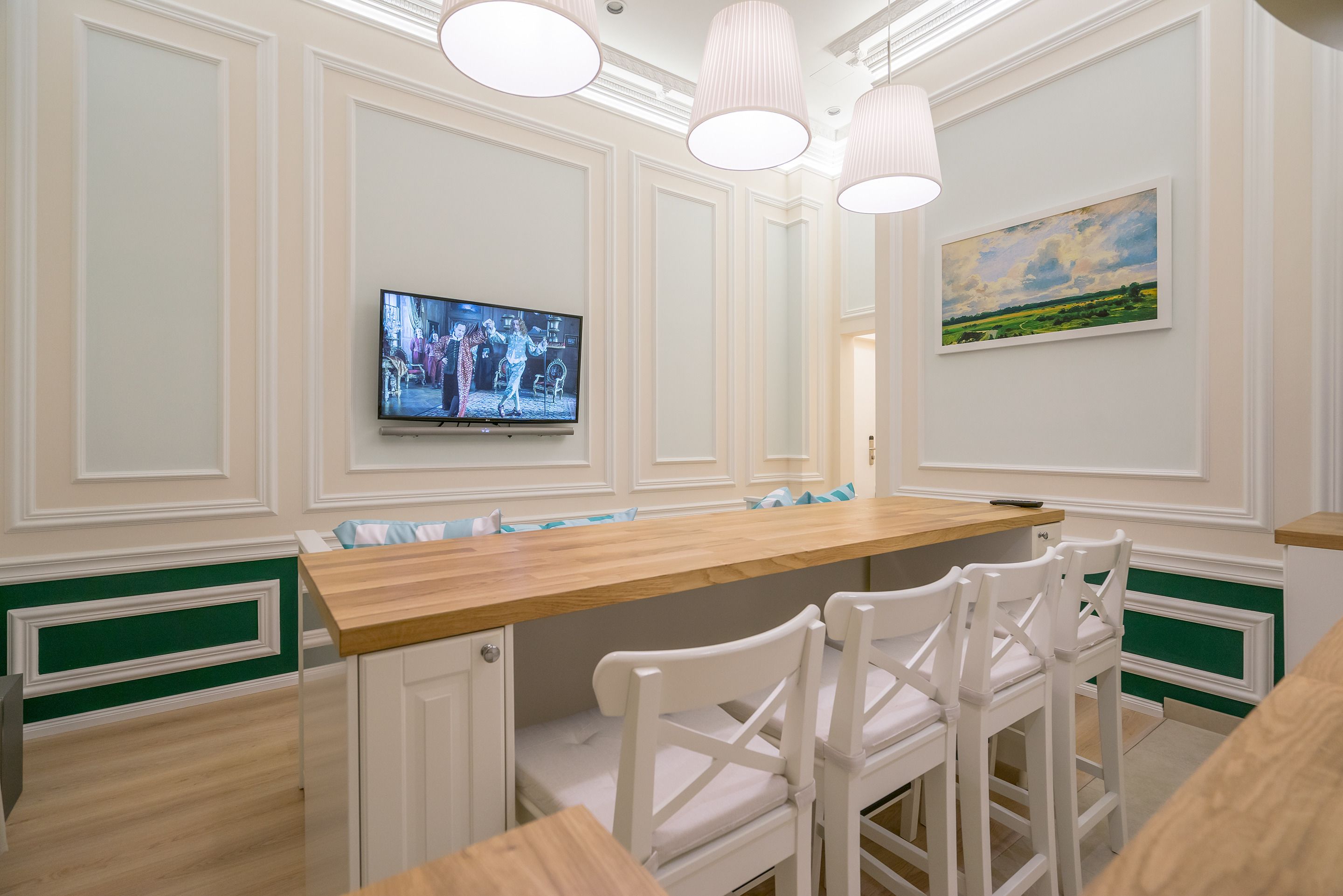 A dining area with a wooden table and white chairs, a television mounted on the wall, and a painting above the table.