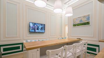 A dining area with a wooden table and white chairs, a television mounted on the wall, and a painting above the table.