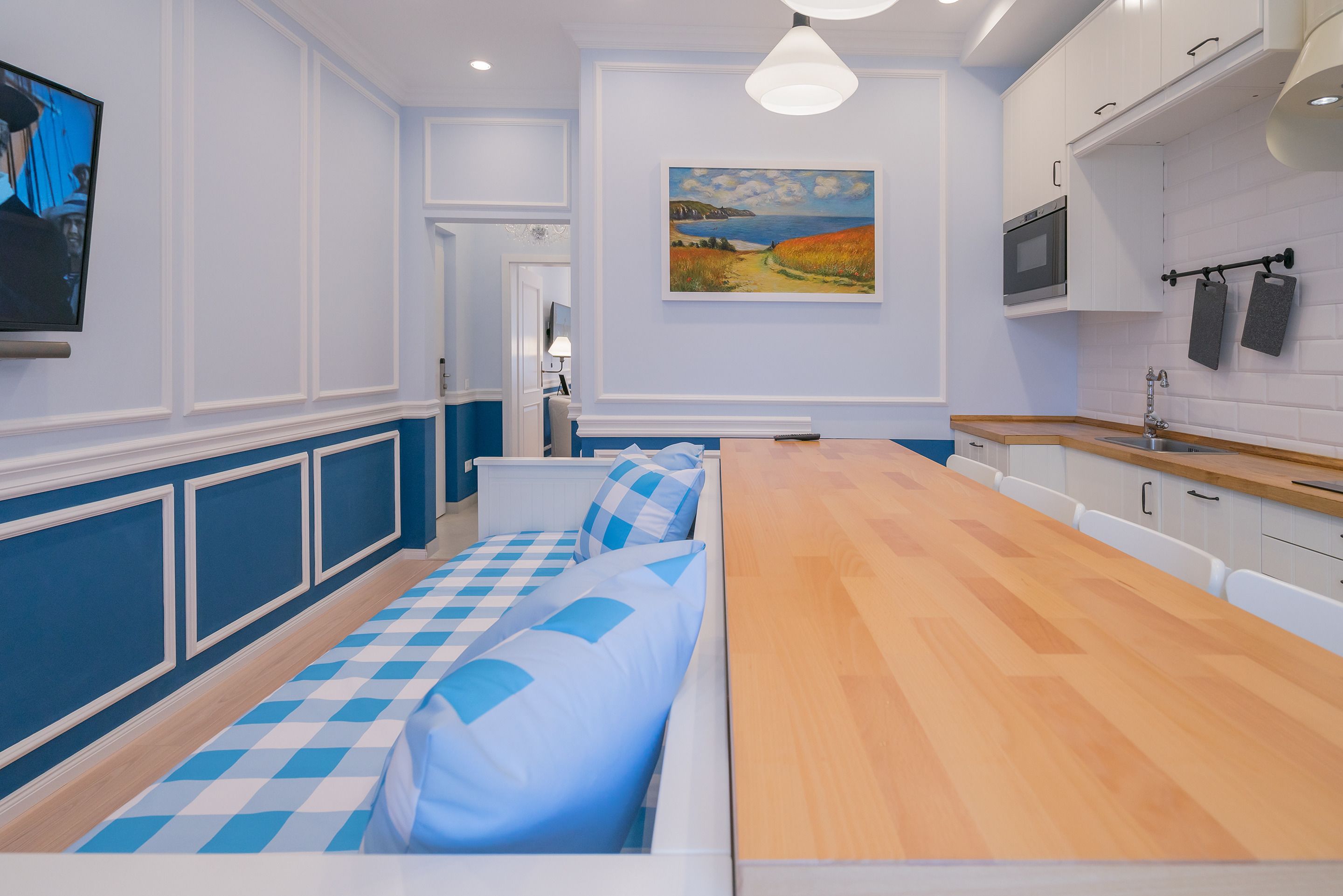 A modern kitchen with a long wooden table, white cabinets, and a painting of a landscape on the wall.