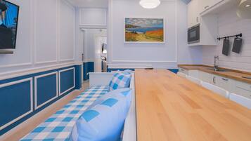 A modern kitchen with a long wooden table, white cabinets, and a painting of a landscape on the wall.