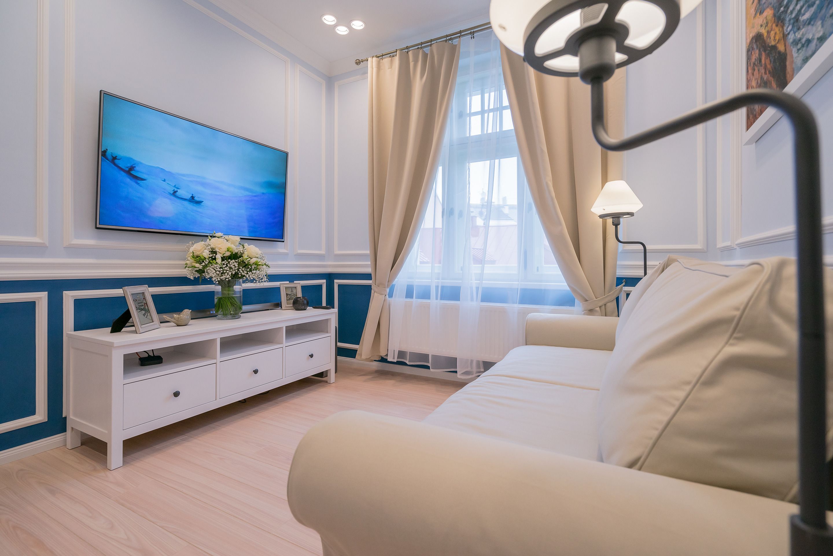 A modern living room with a white sofa, a white TV stand with a painting, and a large window with curtains.