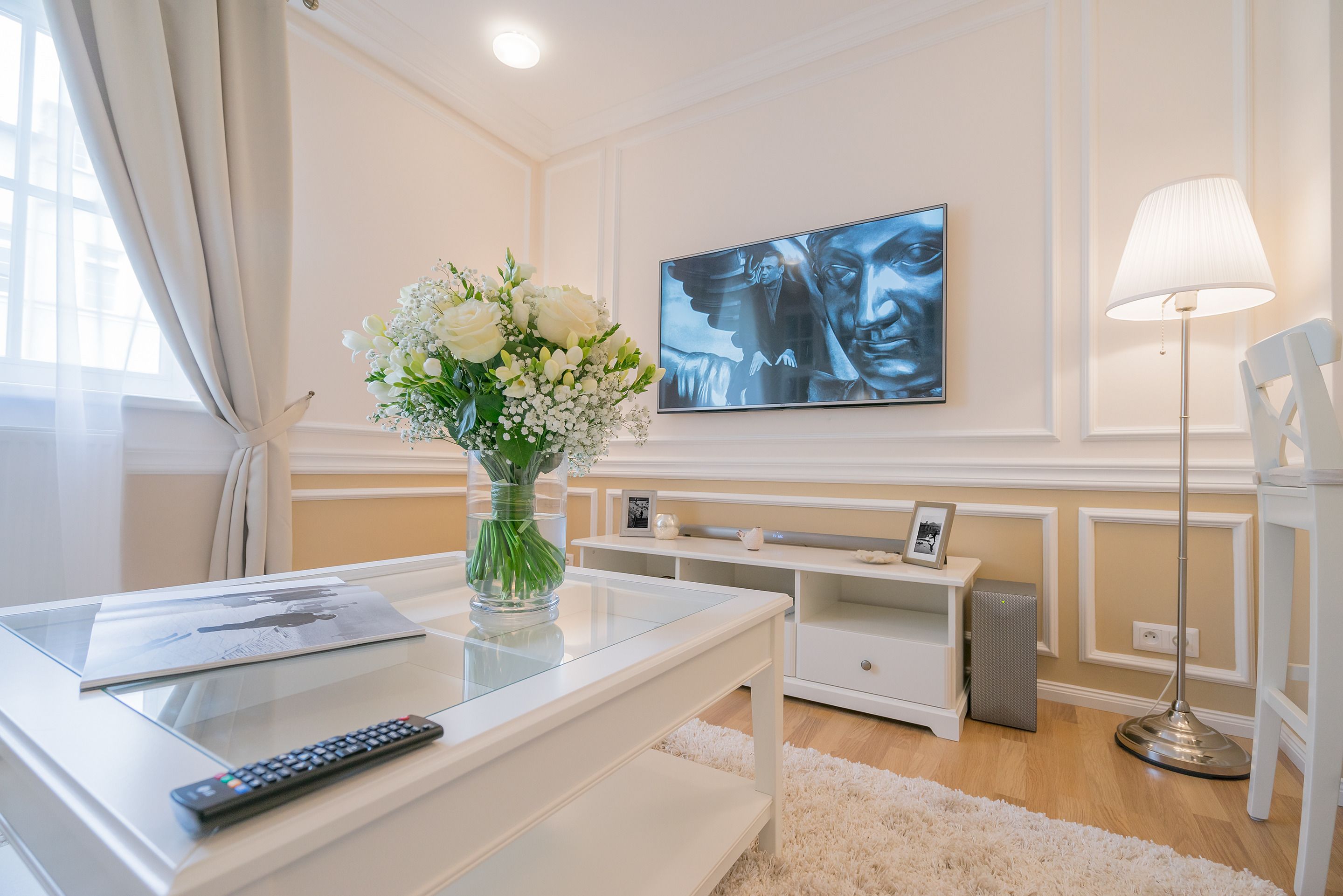 A modern living room with a glass coffee table, a remote control, a vase of flowers, a flat-screen TV, and a standing lamp.