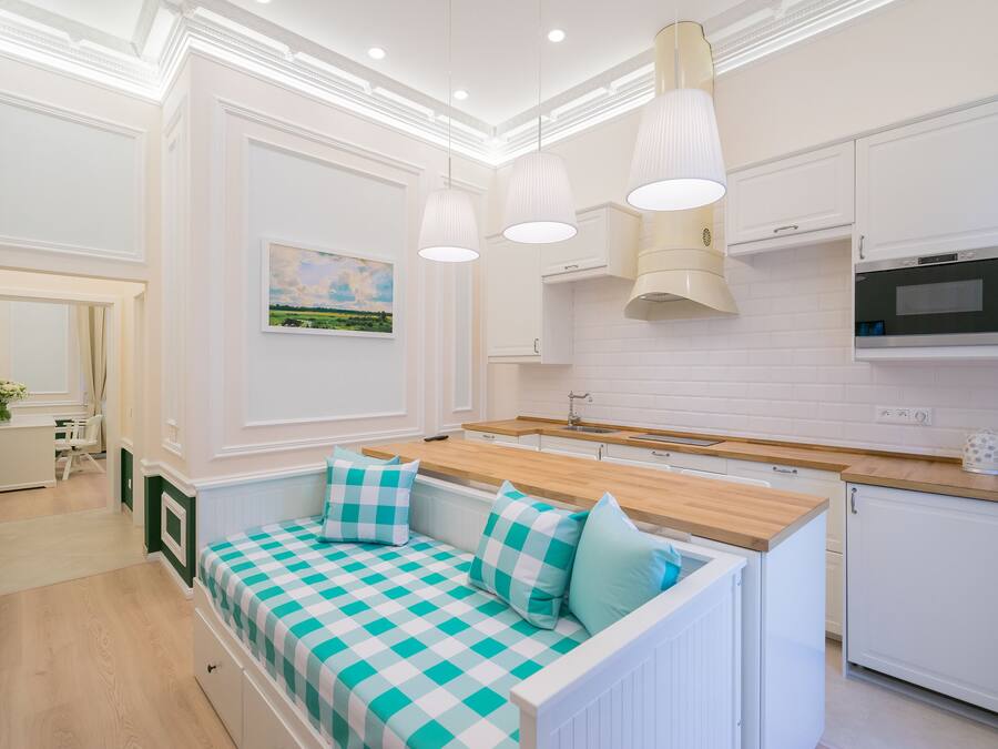A modern kitchen with white cabinetry, a wooden countertop, and a built-in oven. There is a seating area with a checkered bench and cushions.