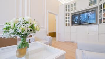 Een moderne woonkamer met een glazen salontafel, witte banken, een televisie bevestigd aan een witte kast, en een bloevas op de tafel.