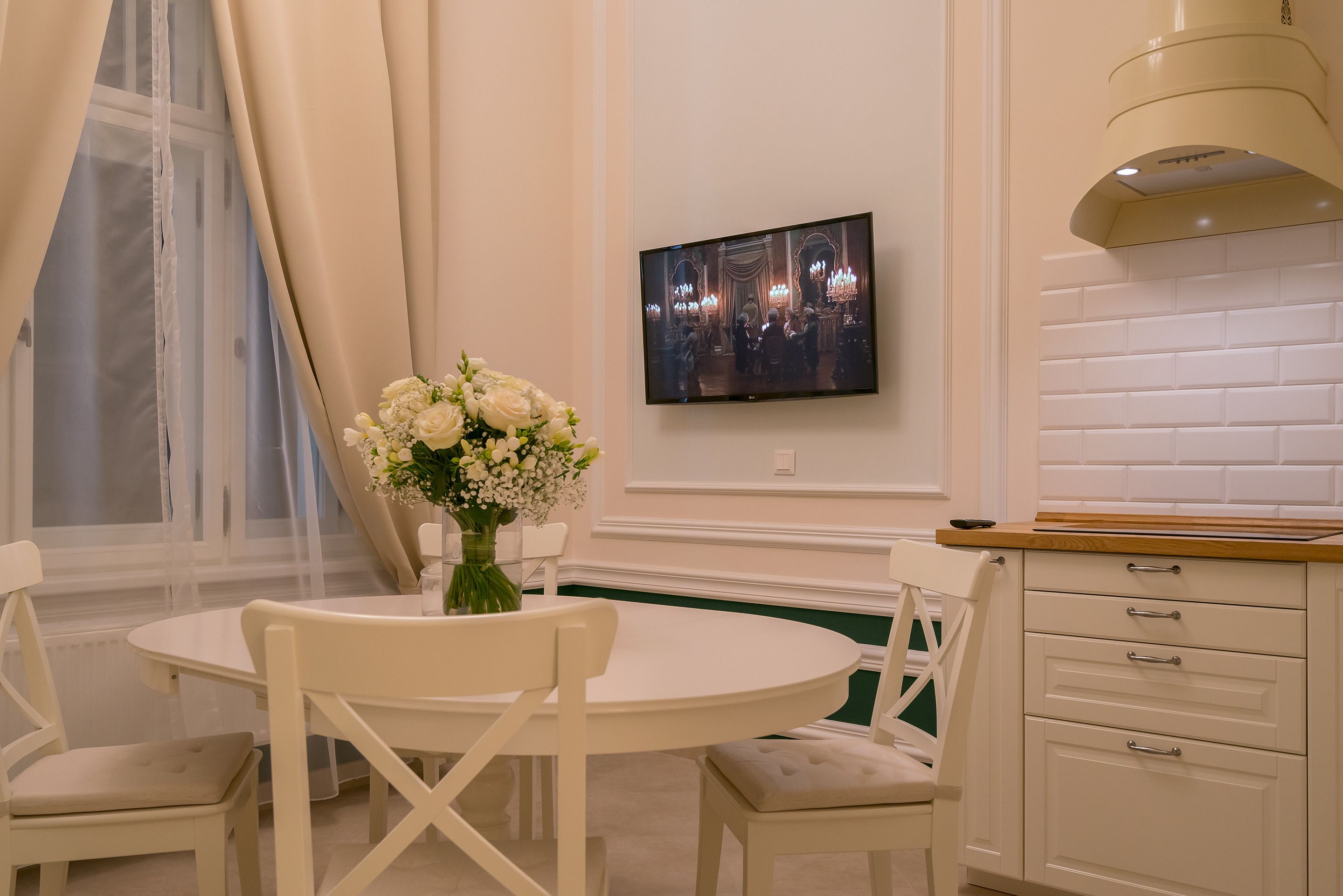 A dining area with a round table, white chairs, a vase of flowers, and a television mounted on the wall.