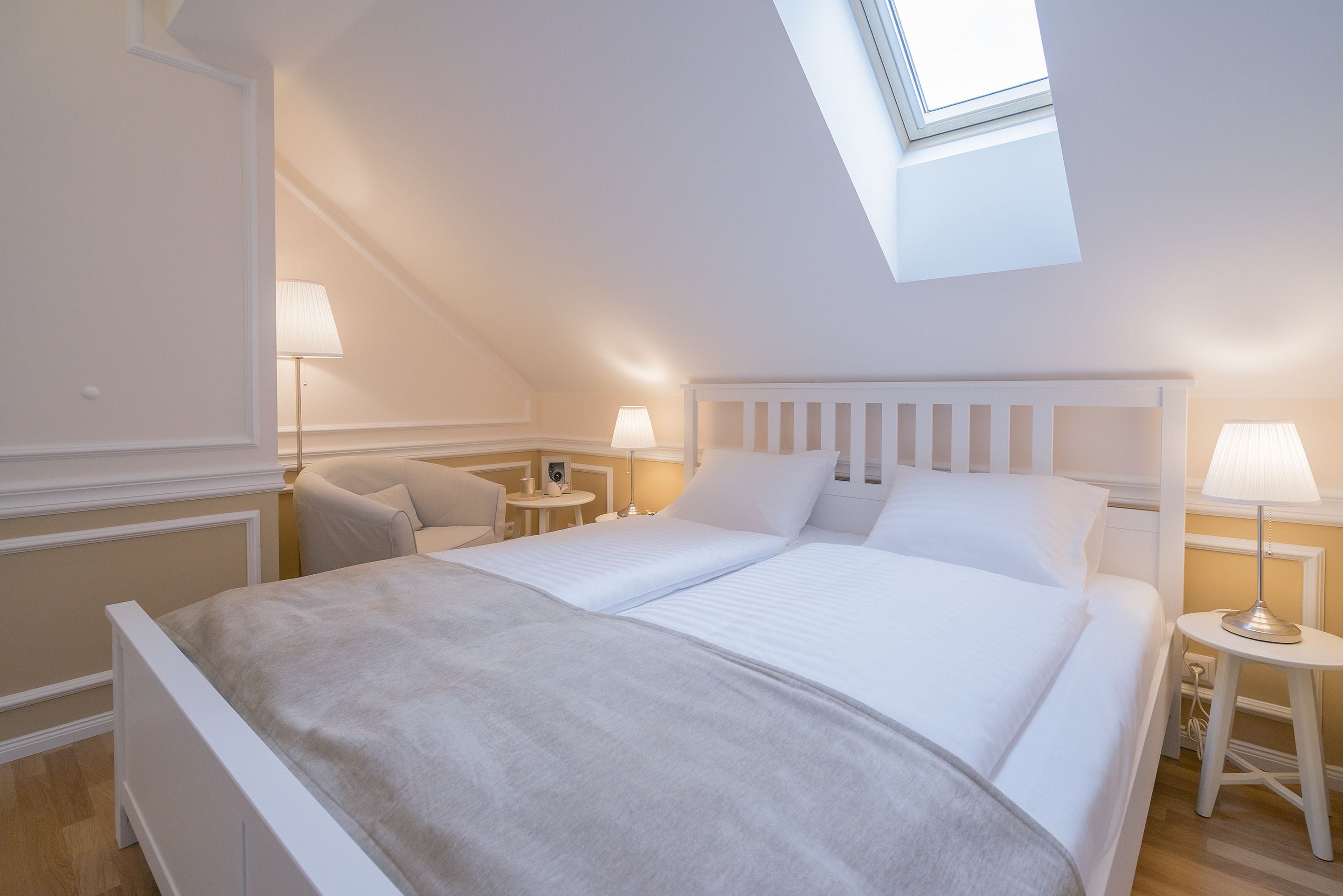 A bedroom with a skylight, a bed with white bedding, two bedside tables with lamps, and a chair.
