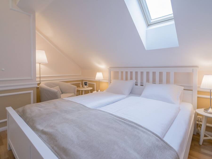 A bedroom with a skylight, a bed with white bedding, two bedside tables with lamps, and a chair.