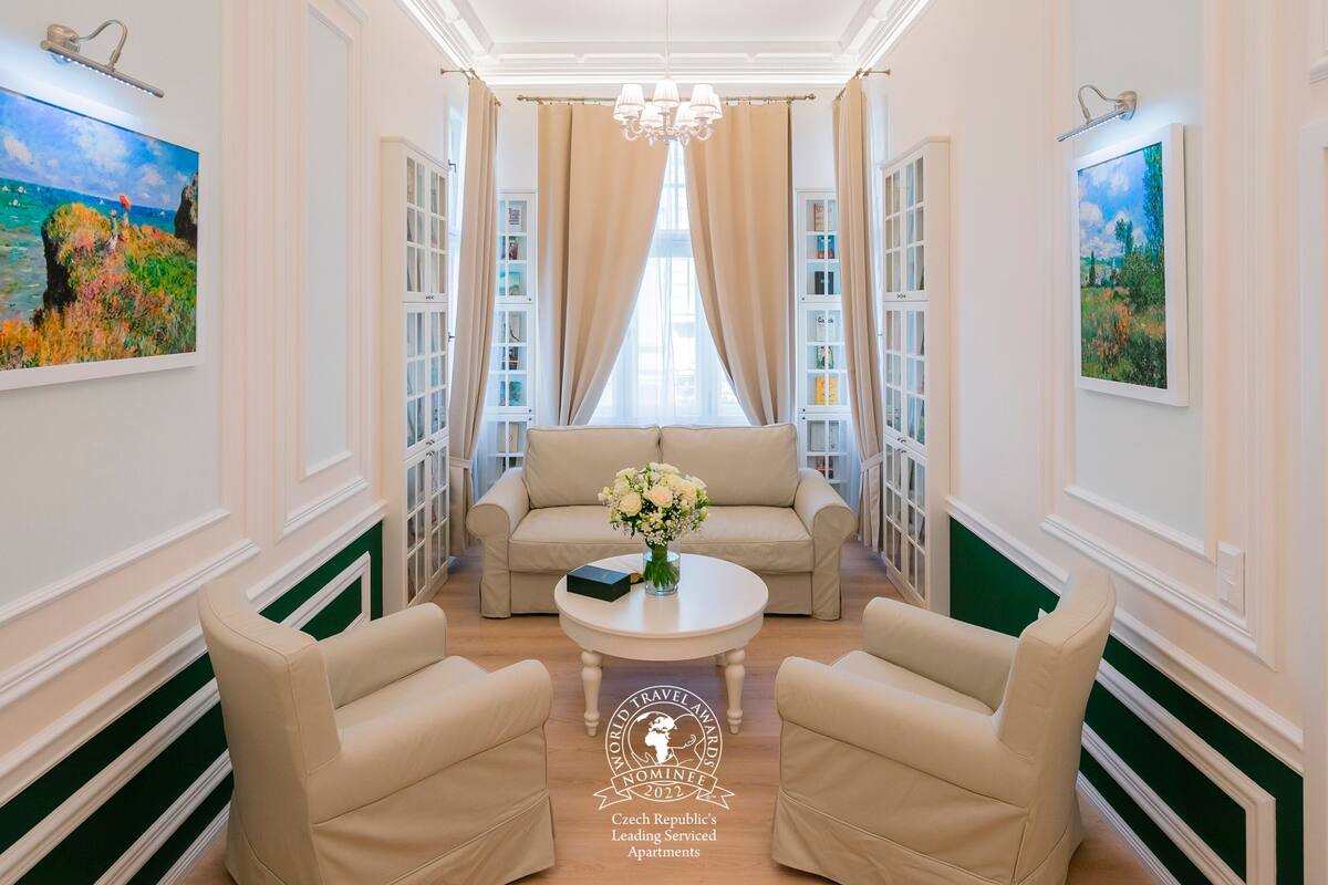 A well-lit, elegantly furnished living room with a sofa, two armchairs, a coffee table with a vase of flowers, and a bookshelf.