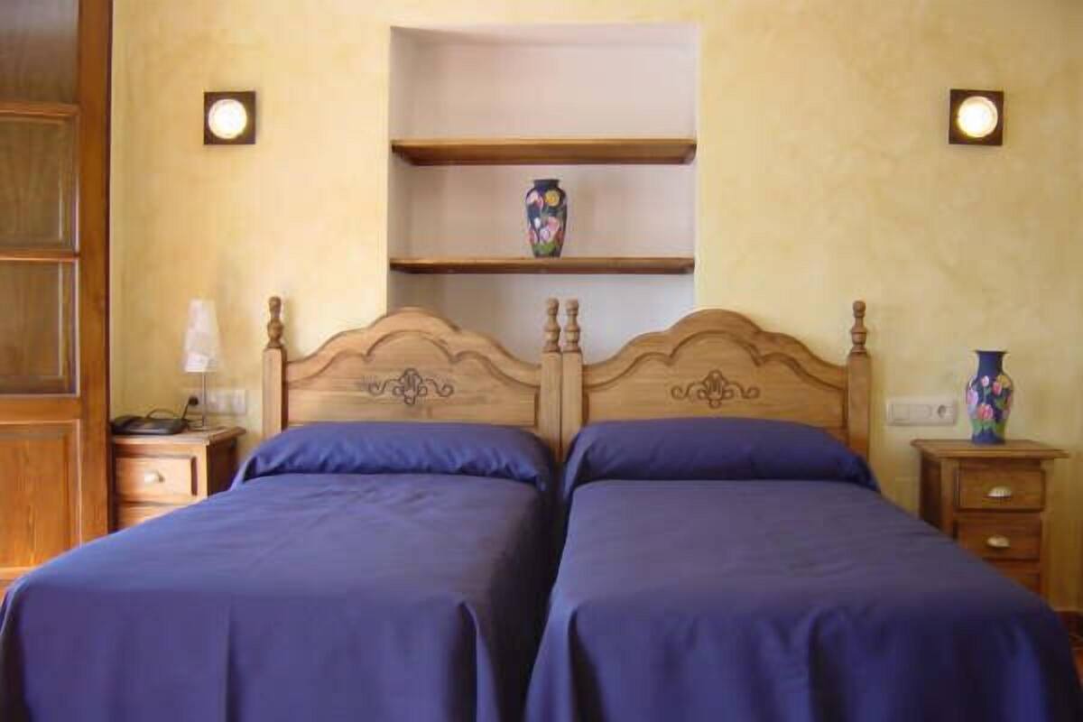 A room with two beds, wooden headboards, a shelf with a vase, and bedside tables with lamps.