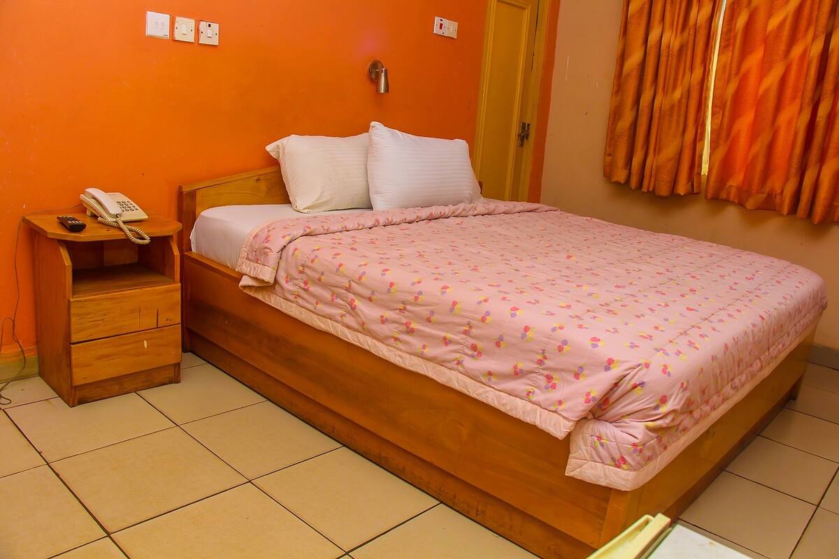 A single bed with a pink floral bedspread, a wooden nightstand with a telephone, and a wall socket.