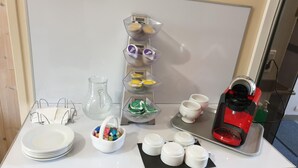 Frühstücksaufstellung mit Kaffeemaschine, Tassen, Tellern und einer Schüssel mit bunten Gegenständen auf einer Arbeitsplatte.