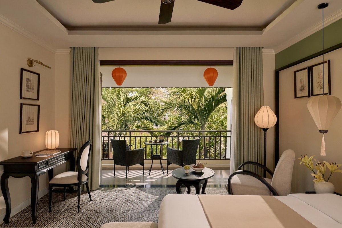 A hotel room with a balcony, a desk, two chairs, a bed, and a table with a vase of flowers.