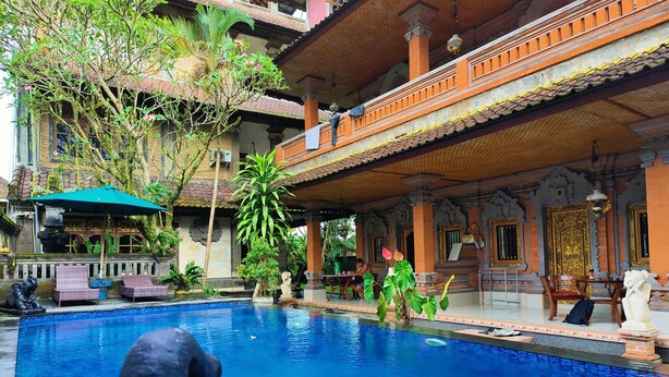 Una piscina con una estatua en primer plano, rodeada por un edificio tradicional de arquitectura ornamentada y una zona de estar cubierta.