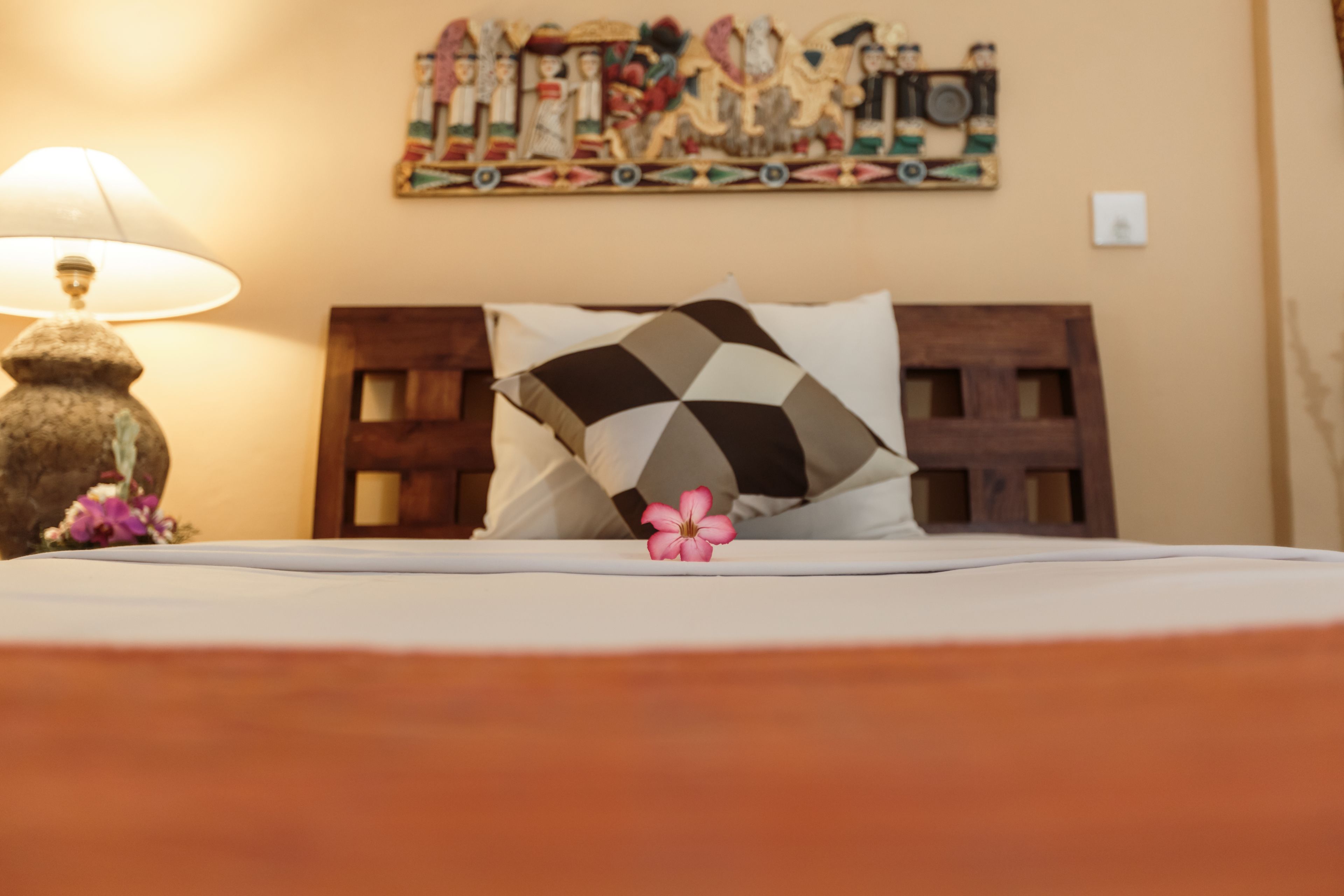 A bed with a geometric patterned pillow, a pink flower, and a lamp on a wooden nightstand.
