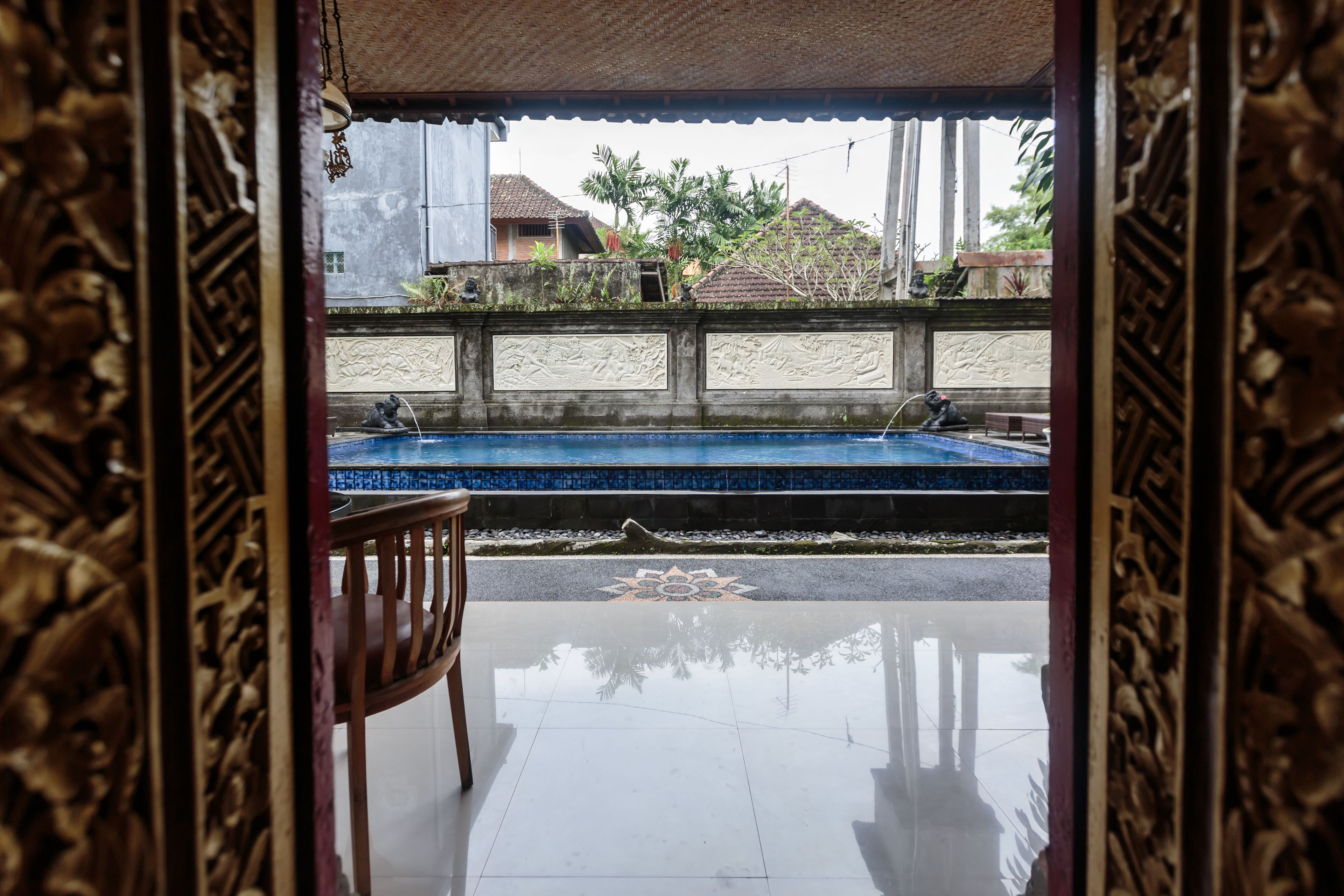Vista de una piscina con una silla de madera en primer plano, un muro decorativo y, al fondo, casas y árboles.