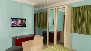 A room with teal walls, a television mounted on the wall, a beige armchair, a red cabinet, and a sliding glass door with green curtains. - Divine Foutain Apapa Branch (Lagos)