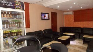A hotel room with a seating area featuring black leather sofas and small tables, a minibar with various beverages, and a television mounted on the wall. - Divine Foutain Apapa Branch (Lagos)