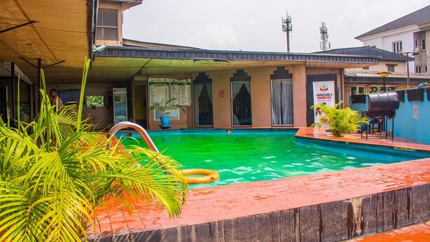 A swimming pool with a wooden deck and palm plants, surrounded by buildings with a sign for a barbecue restaurant. - Divine Fountain Agidingbi (Lagos)