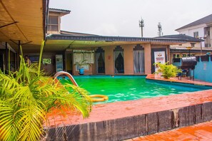 A swimming pool with a wooden deck and palm plants, surrounded by buildings with a sign for a barbecue restaurant. - Divine Fountain Agidingbi (Lagos)