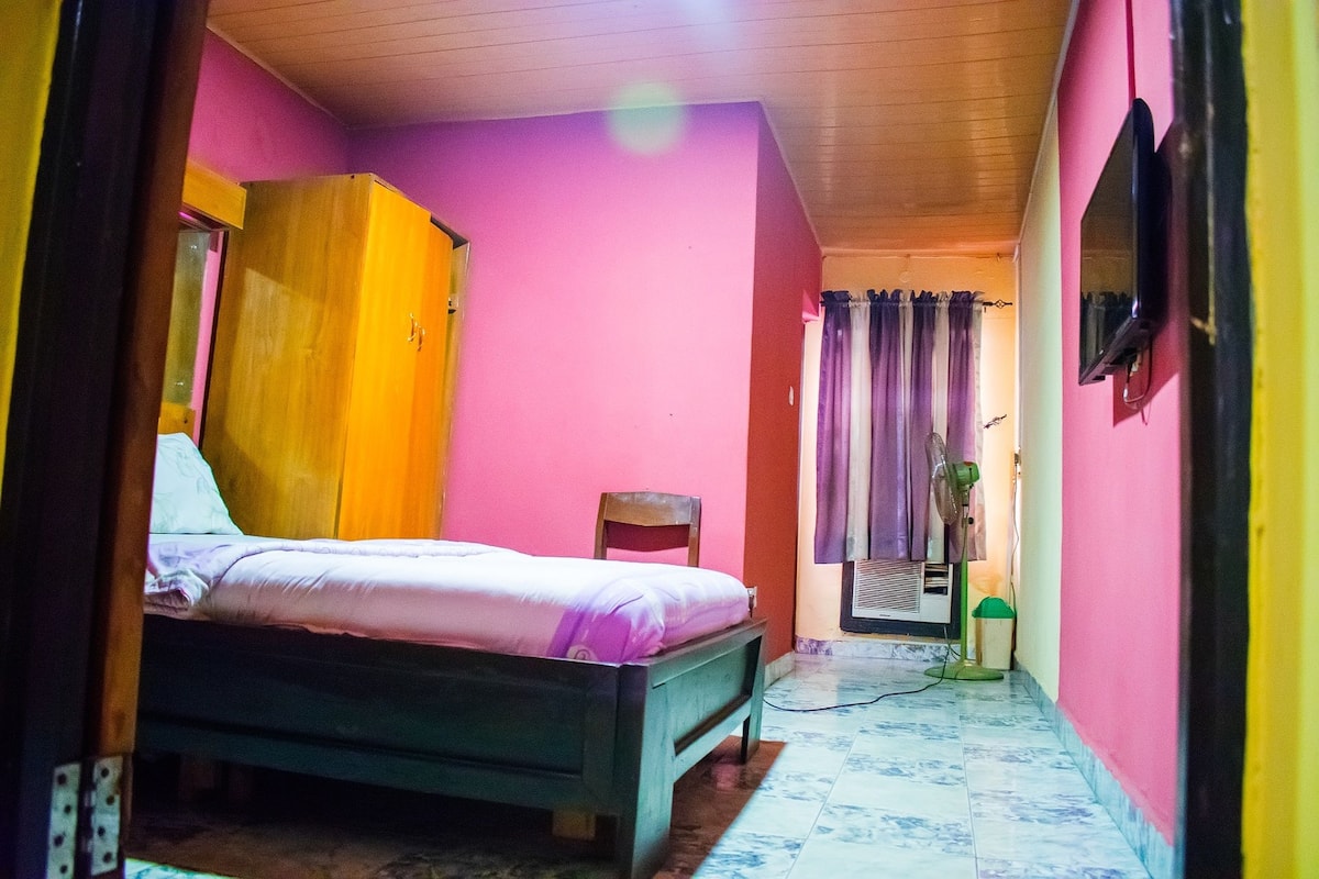 A bedroom with a bed, a wooden wardrobe, a chair, and a television on the wall.