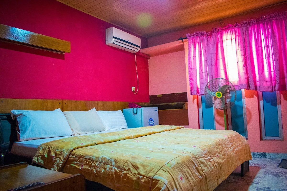 A room with a bed, an air conditioner, a fan, and a window with curtains.