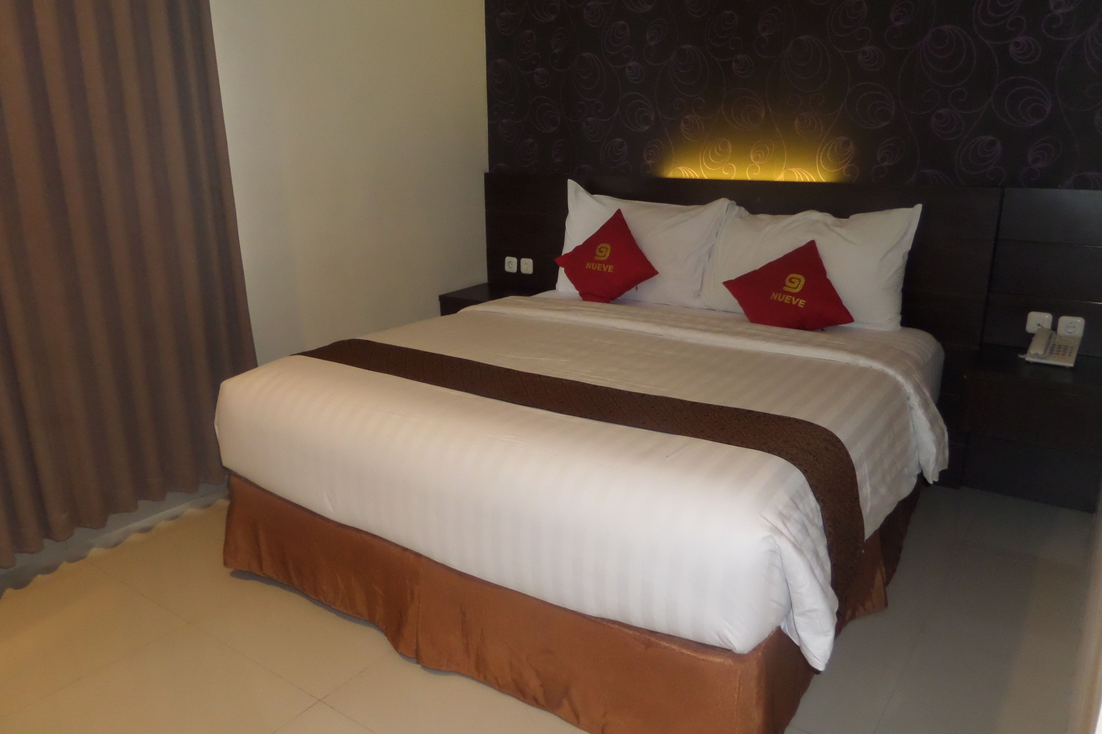 A hotel room with a large bed, a headboard with a light fixture, and a dark patterned wall.