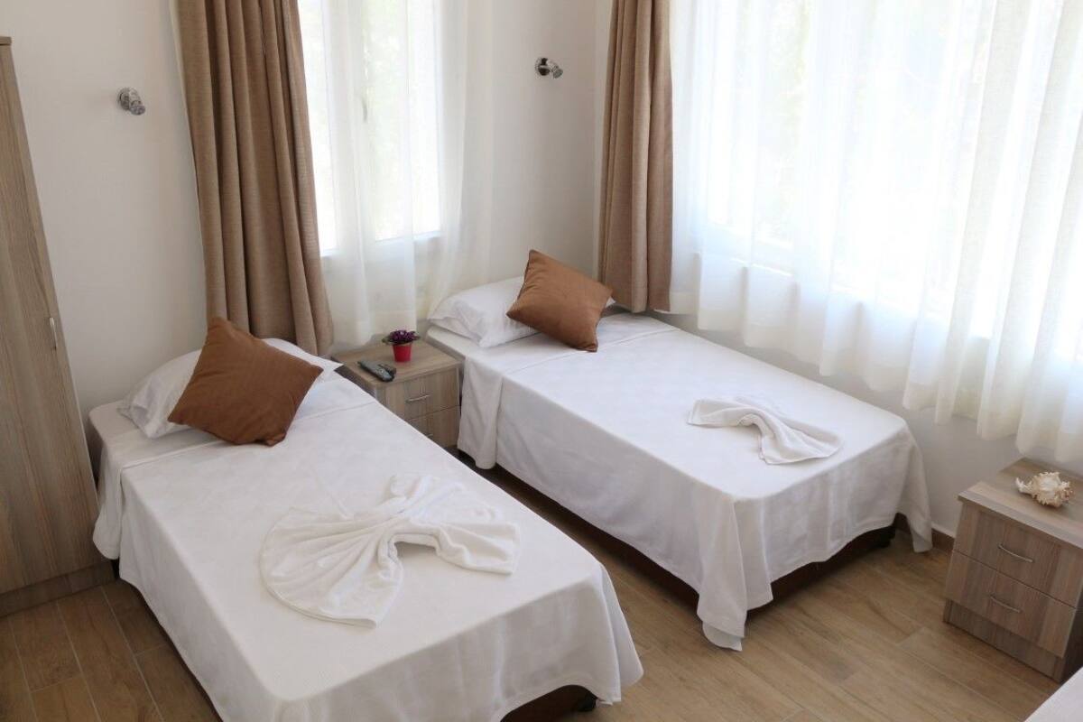 Habitación de hotel con dos camas individuales, cada una con cubrecama blanco y almohada marrón. Se aprecia un mesita de noche de madera con una taza roja y un pequeño objeto decorativo. Las paredes son de color claro y el suelo es de madera.