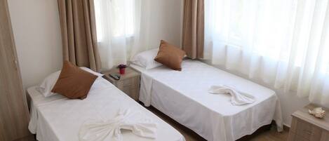 A hotel room with two single beds, each with a white bedspread and a brown pillow. There is a wooden nightstand with a red cup and a small decorative item on it. The room has light-colored walls and a wooden floor.