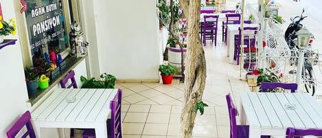An outdoor café with white tables and purple chairs, surrounded by potted plants and a tree.