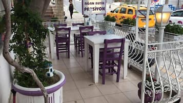 An outdoor seating area with tables and chairs, a potted plant, and a street view with a taxi and a pedestrian.