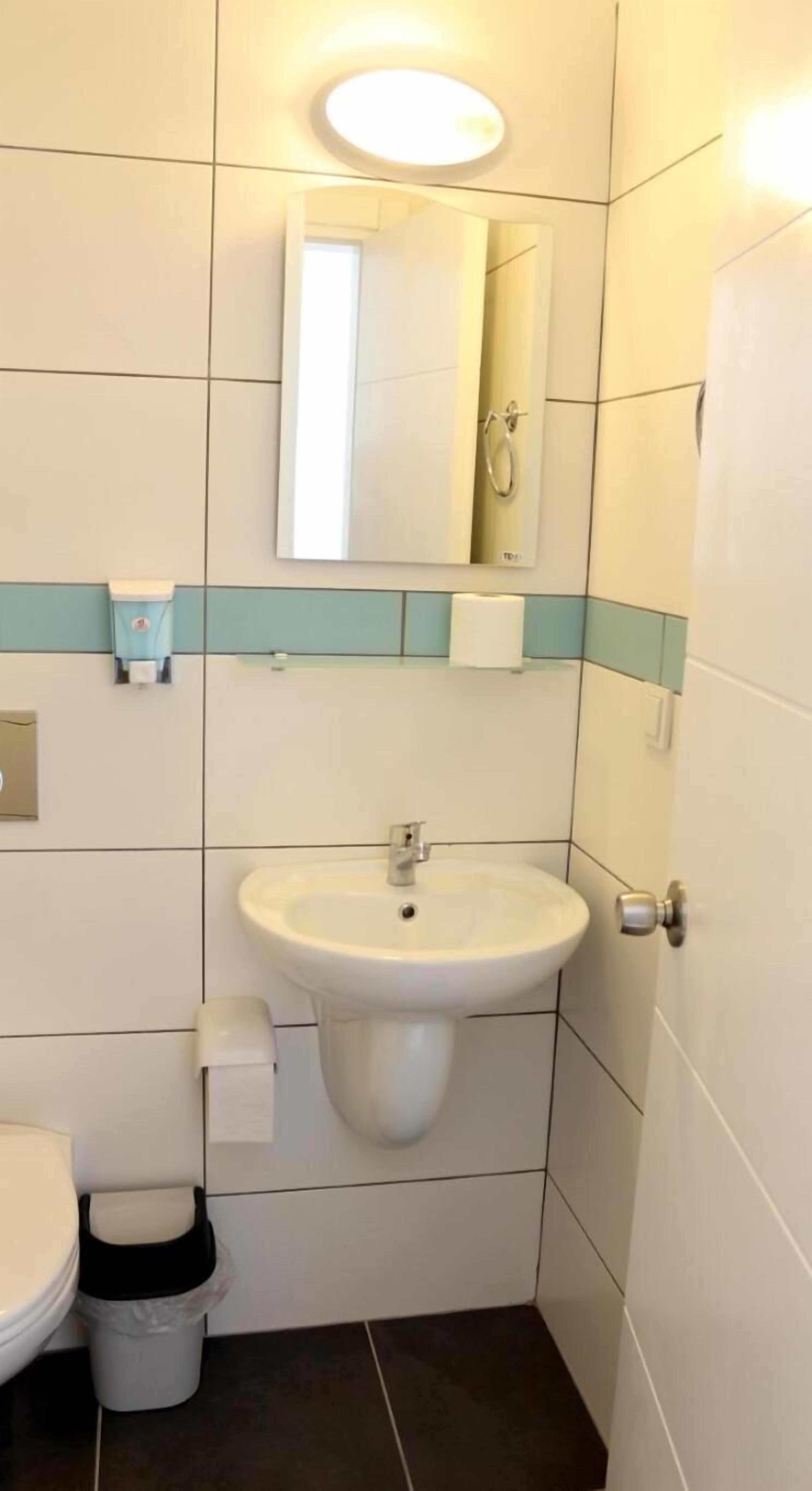 Een compacte badkamer met een witte wastafel, een spiegel en een toilet. Ook een prullenbak en een handdoekdispenser zijn aanwezig.