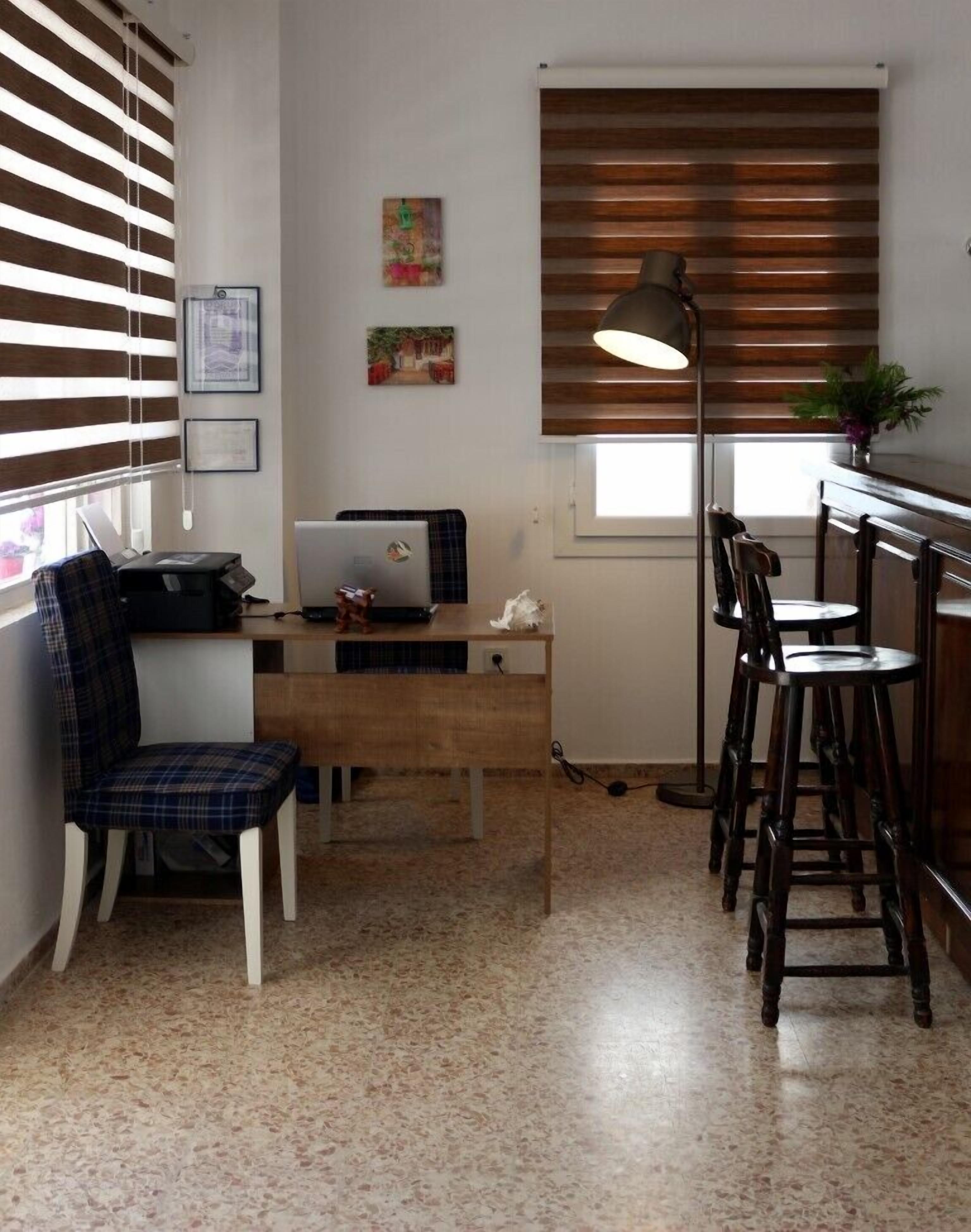 A compact living space with a wooden desk, a laptop, a plaid chair, a bar stool, and a floor lamp.