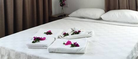 A hotel room with a bed, white linens, brown curtains, an air conditioner, and a vase with flowers on the nightstand.