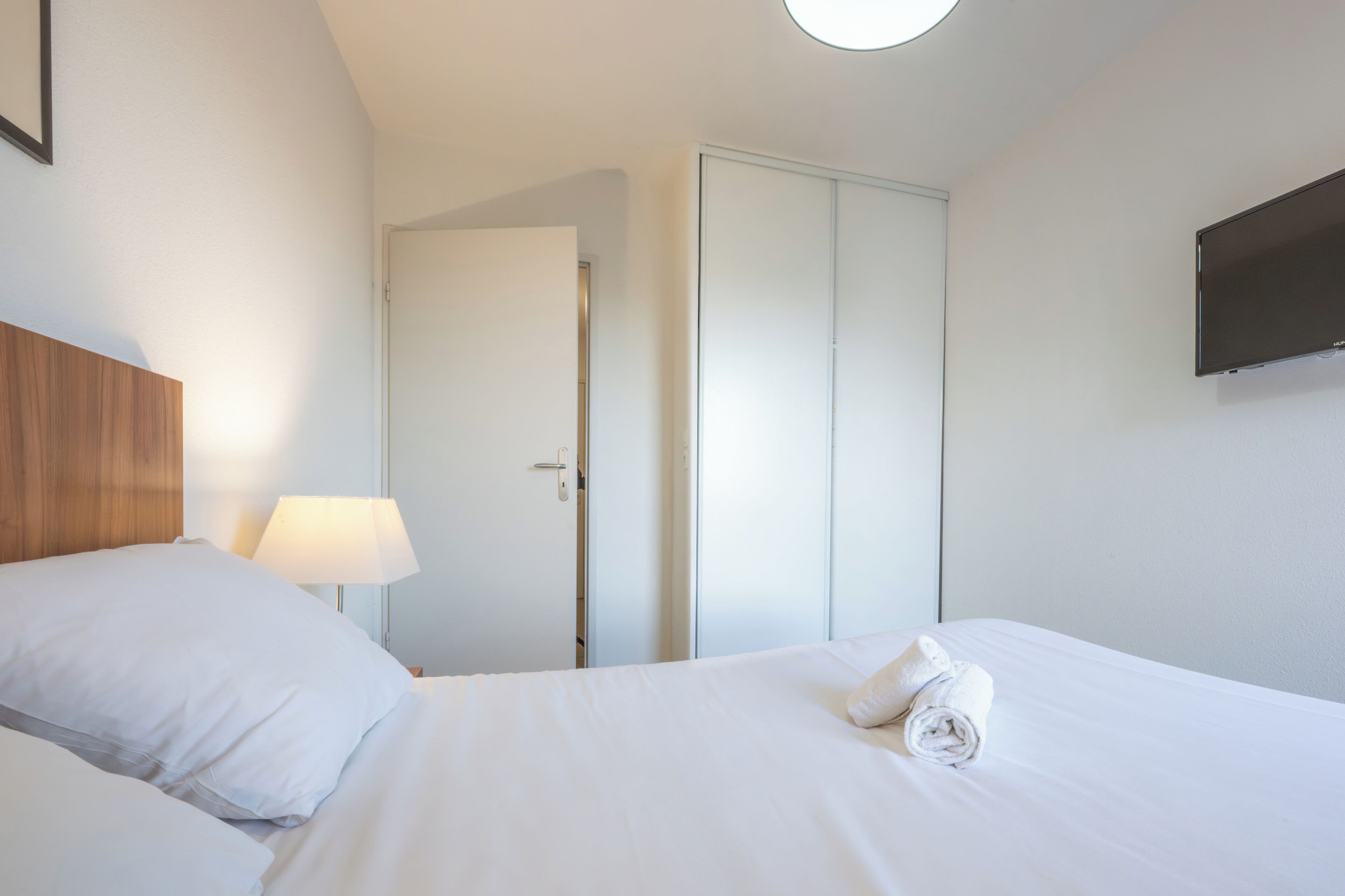 Een hotelkamer met een bed, een nachtkastje met een lamp, een kledingkast en een flatscreen televisie aan de muur.