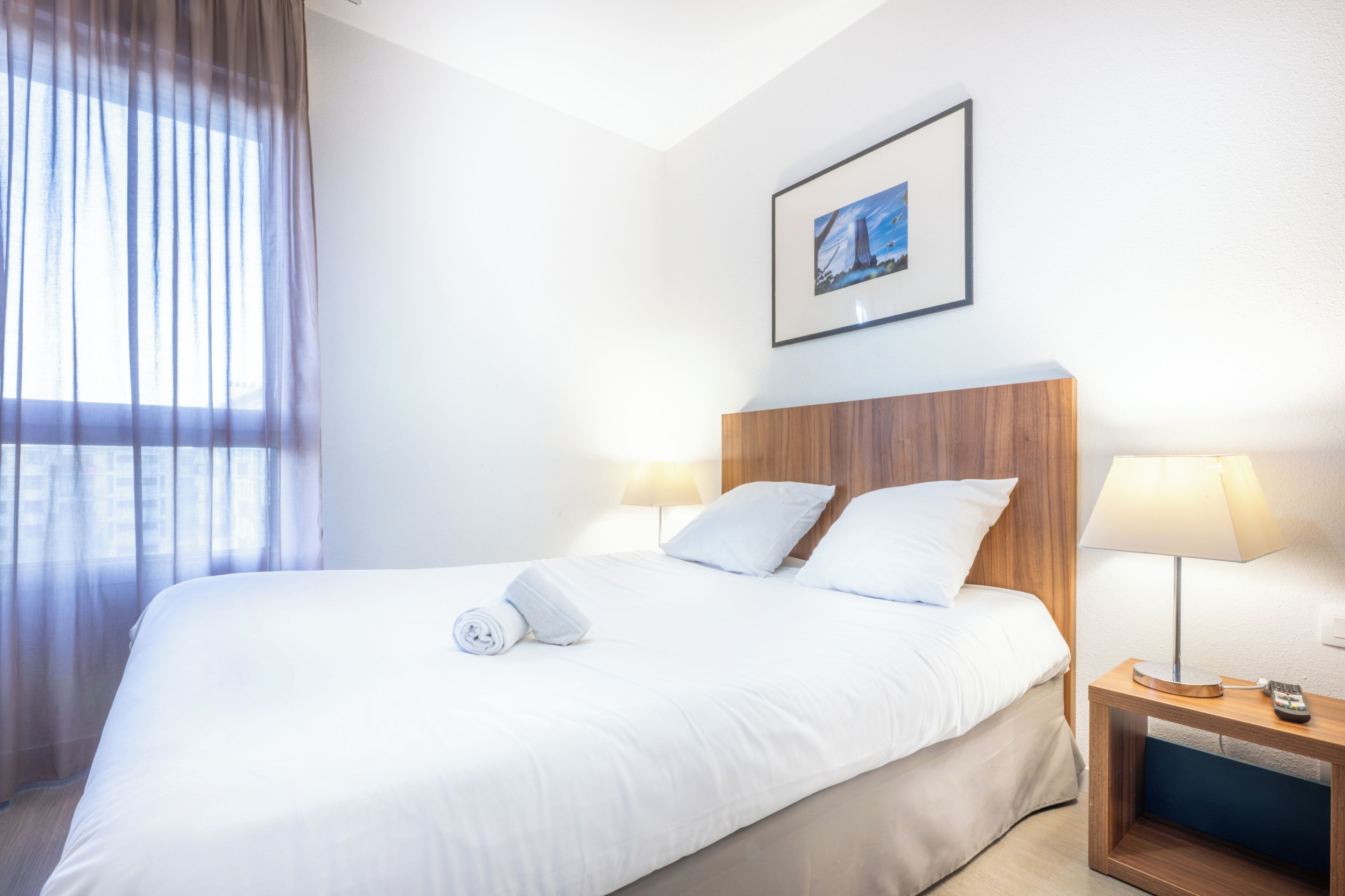 Un letto rifatto con lenzuola bianche, una testiera in legno, due lampade ai comodini e un quadro appeso al muro.