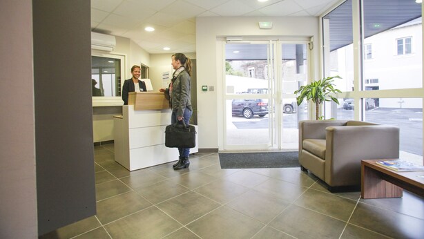 A reception area with a reception desk, a seating area with a bench, and a large window providing a view of the outside. - Appart'hôtel Odalys City - Orléans Centre (Orléans)